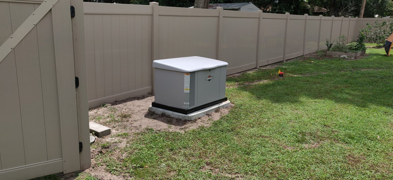 Standby generator installed on a concrete pad beside a vinyl privacy fence in a backyard.